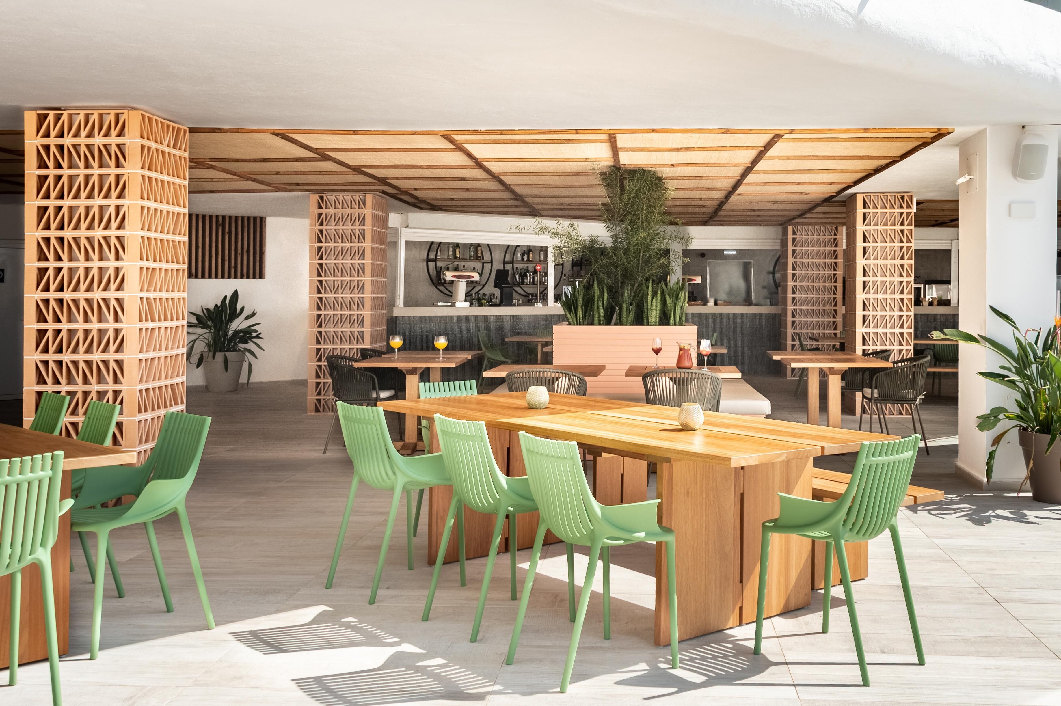 Un espacio para comer al aire libre con mesas de madera y sillas verdes, una barra al fondo y una estructura de techo de madera.