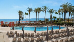 Un espace piscine aménagé avec des chaises longues et des parasols, des palmiers et une vue sur la mer.