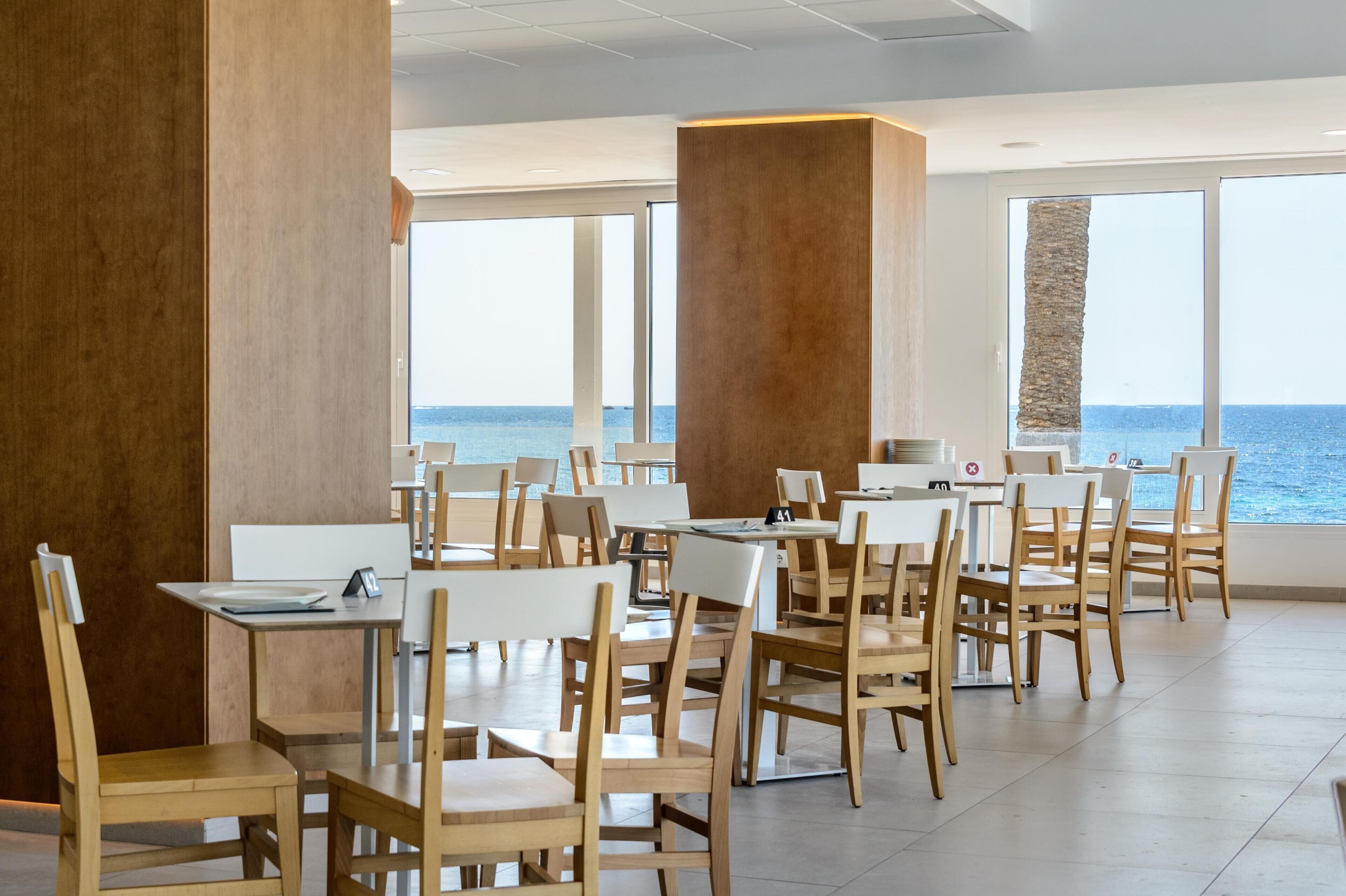 A modern dining area with wooden tables and chairs, large windows offering a view of the sea, and a stone pillar.