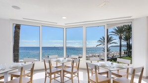 A dining area with large windows overlooking the sea, white furniture, and a view of a beach with umbrellas. - Hotel Vibra Algarb (Ibiza Town)