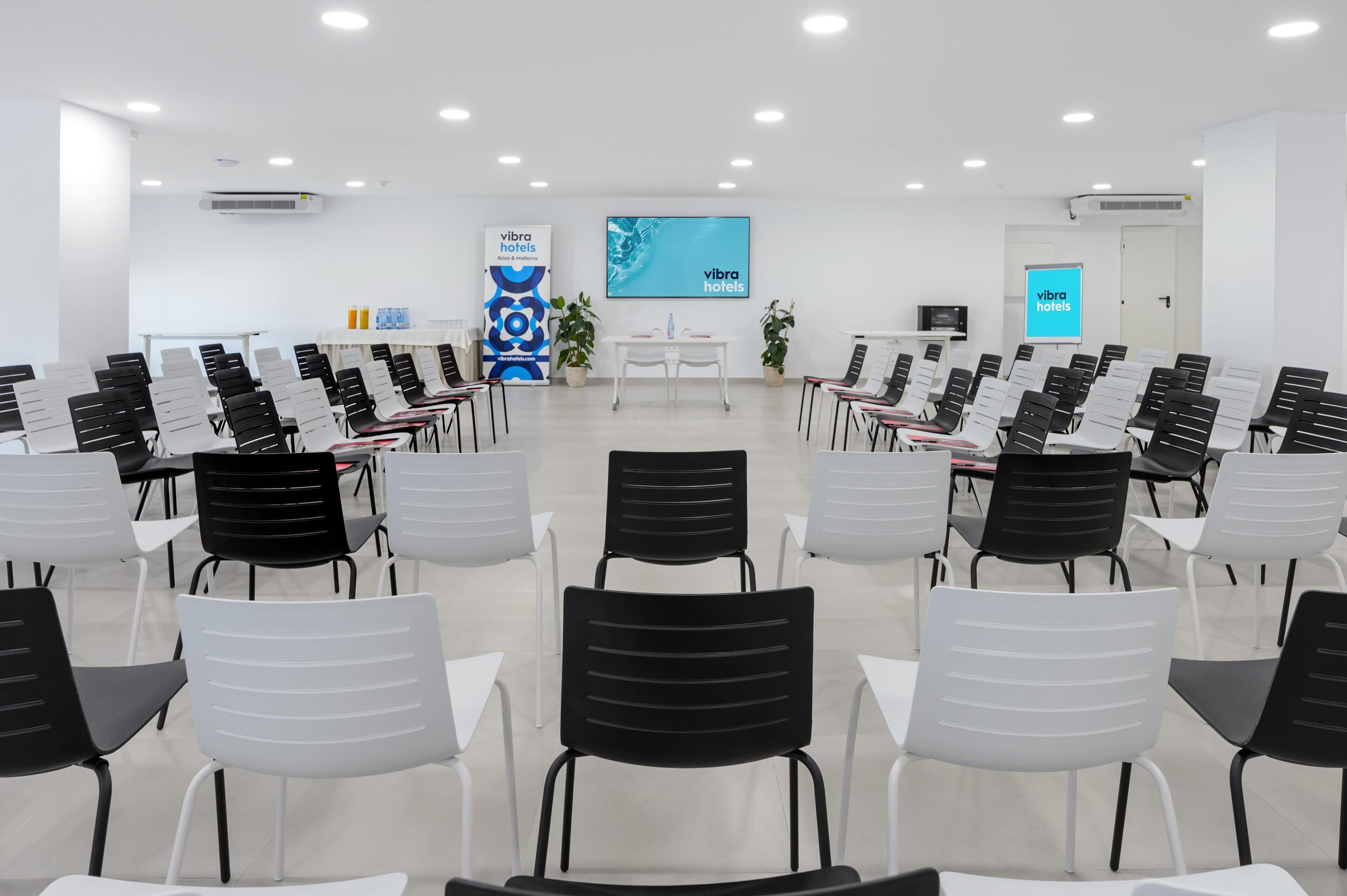 A conference room with rows of chairs, a white table, and a large screen displaying 'vibra hotels'.