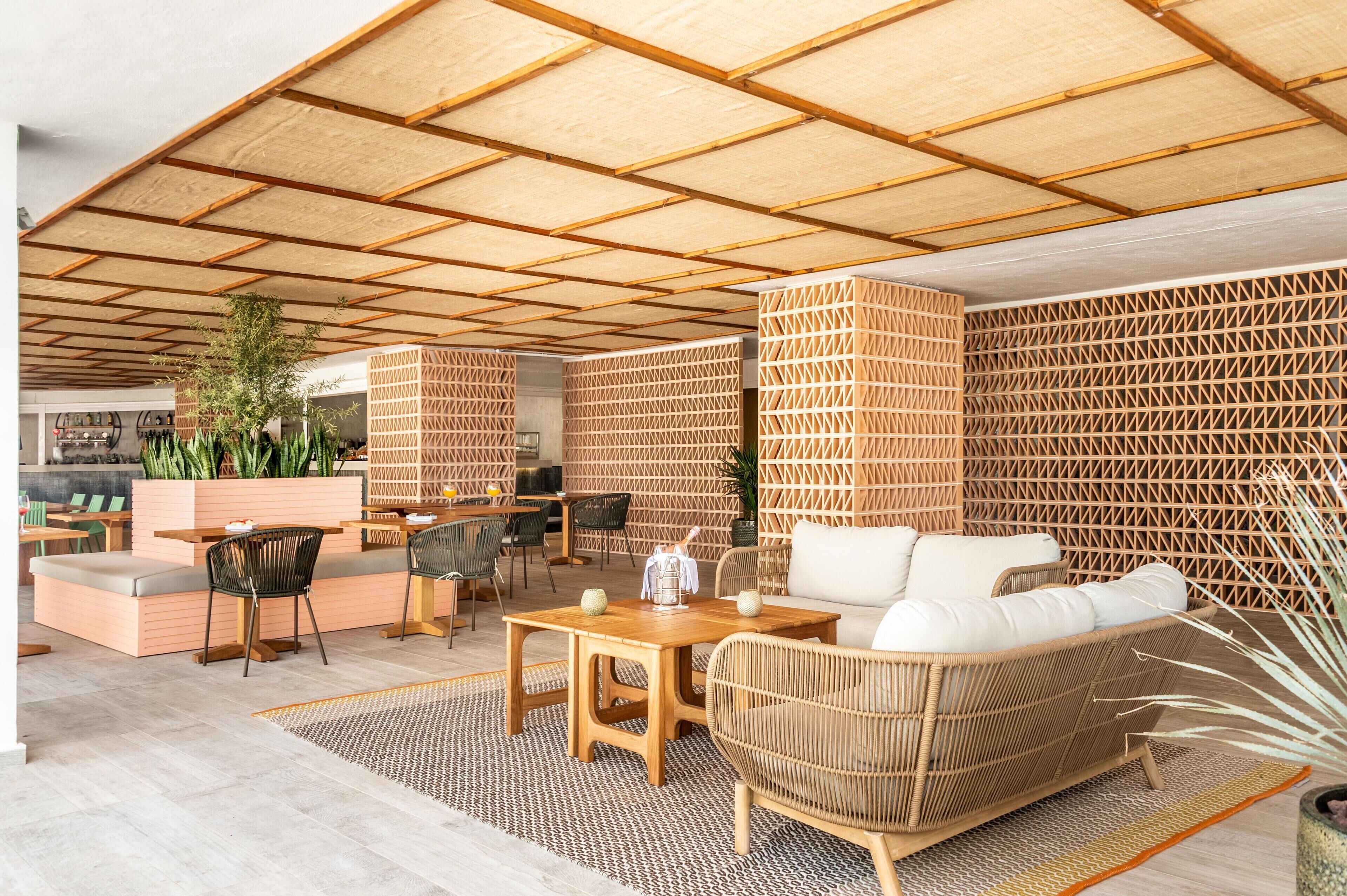 Een terras met houten tafels en stoelen, een rieten bank en een patroonwand.