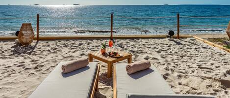Un lounge junto a la playa con tumbonas, una mesa con bebidas y aperitivos, y vista al océano.