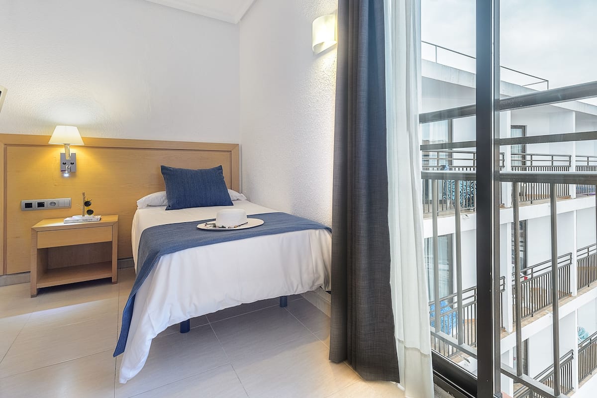 A hotel room with a bed, a nightstand, a lamp, and a view of a building through a window.