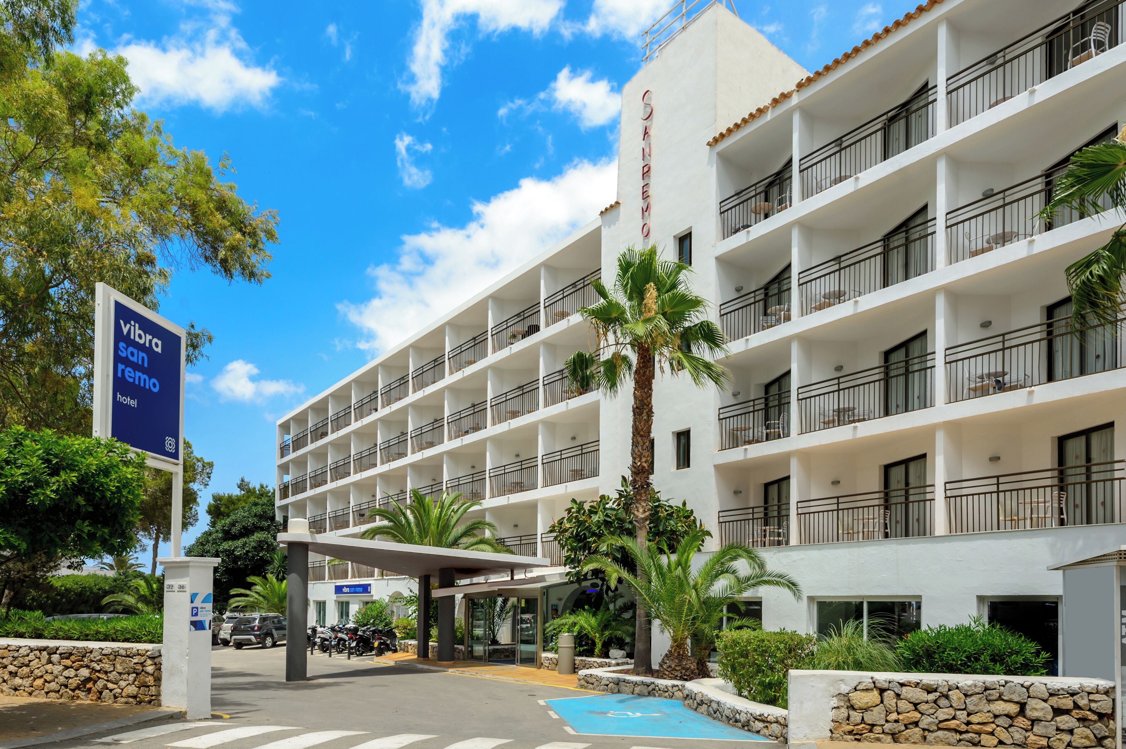 Un hotel de varios pisos con fachada blanca, balcones y una palmera en primer plano.