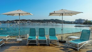 A wooden deck with lounge chairs and umbrellas overlooking a beach with boats and buildings in the background. - Hotel Vibra San Remo (Sant Josep de sa Talaia)