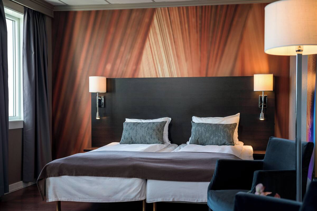 A hotel room with a large bed, two bedside lamps, a dark headboard, and a window with curtains.
