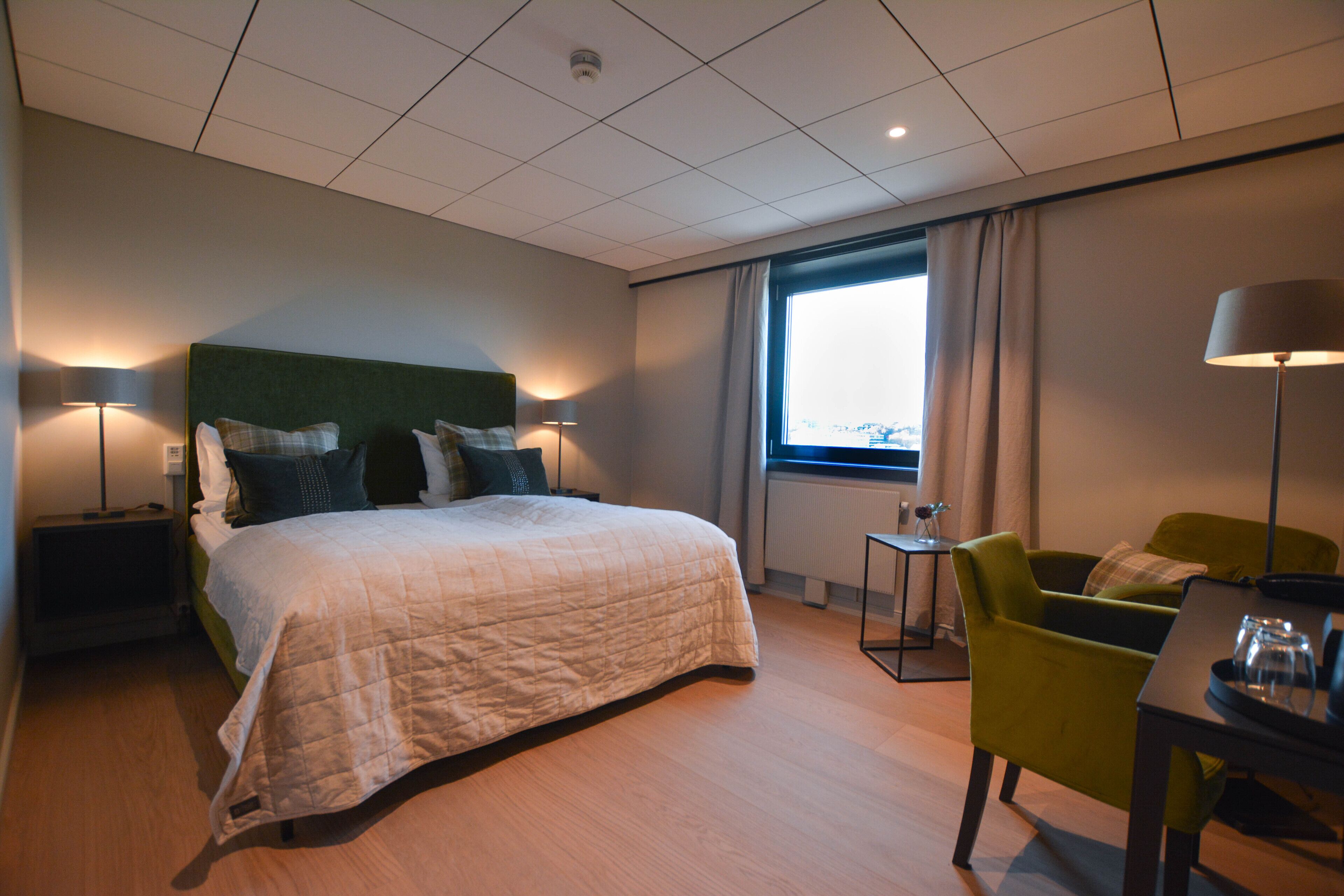 A hotel room with a large bed, a green armchair, a small table, and a window with curtains.