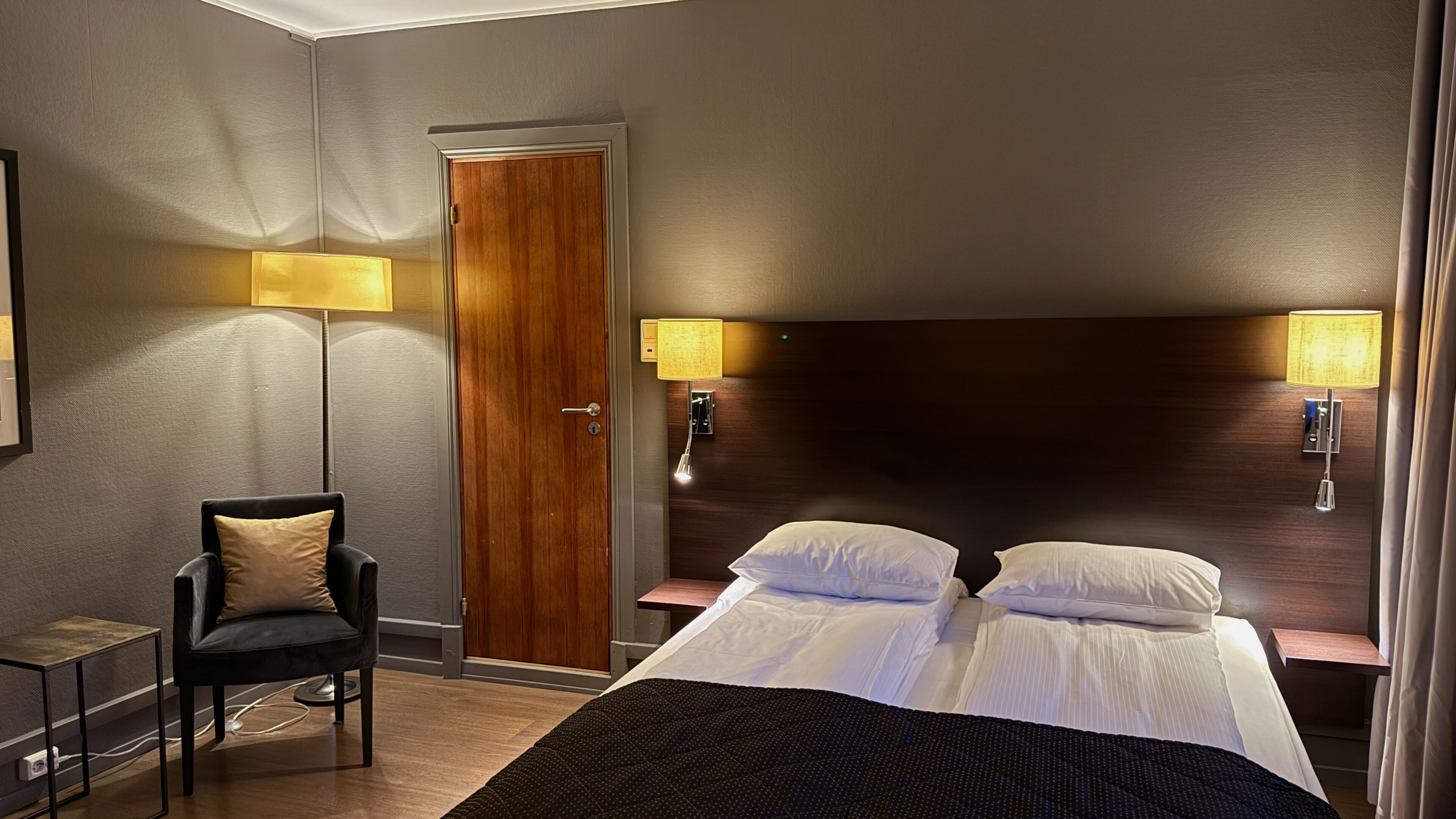 A hotel room with a double bed, two bedside tables with lamps, a wooden door, a small desk with a chair, and a floor lamp.