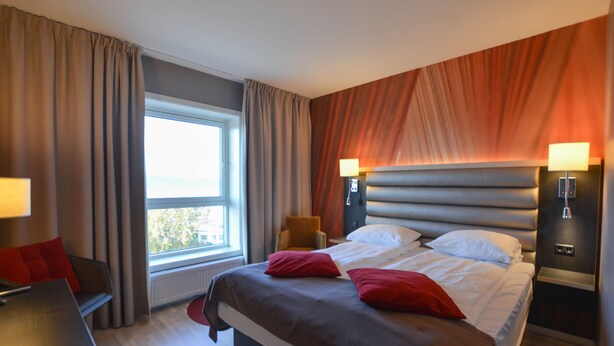 Una habitación de hotel moderna con una cama grande, dos lámparas de noche, una ventana con cortinas, una mesita y una silla.