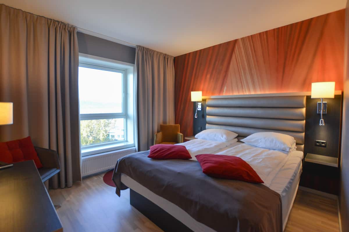 A modern hotel room with a large bed, two bedside lamps, a window with curtains, a small table, and a chair.
