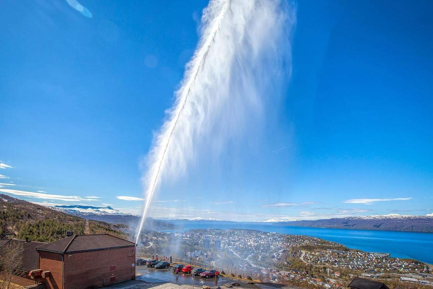A building with a visible water jet feature, a parking area, and a view of a town and lake.