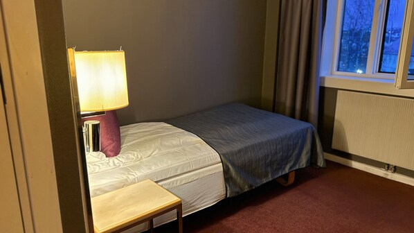 A small, single-bed room with a bedside table, a lamp, a window with curtains, and a radiator. - Quality Hotel Grand Royal (Narvik)