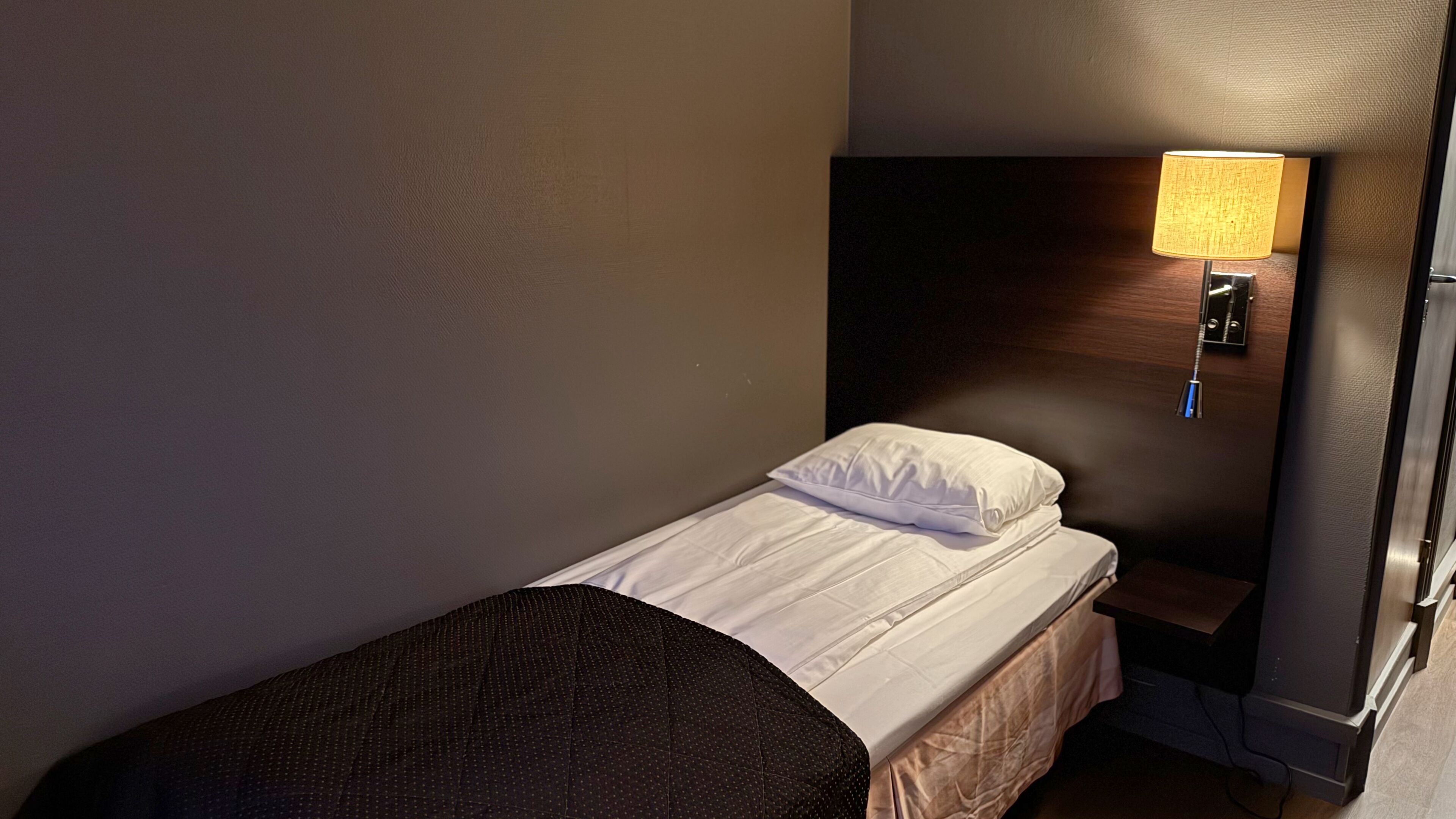 Une petite chambre avec un lit simple, une tête de lit, une lampe de chevet et une table de nuit en bois.