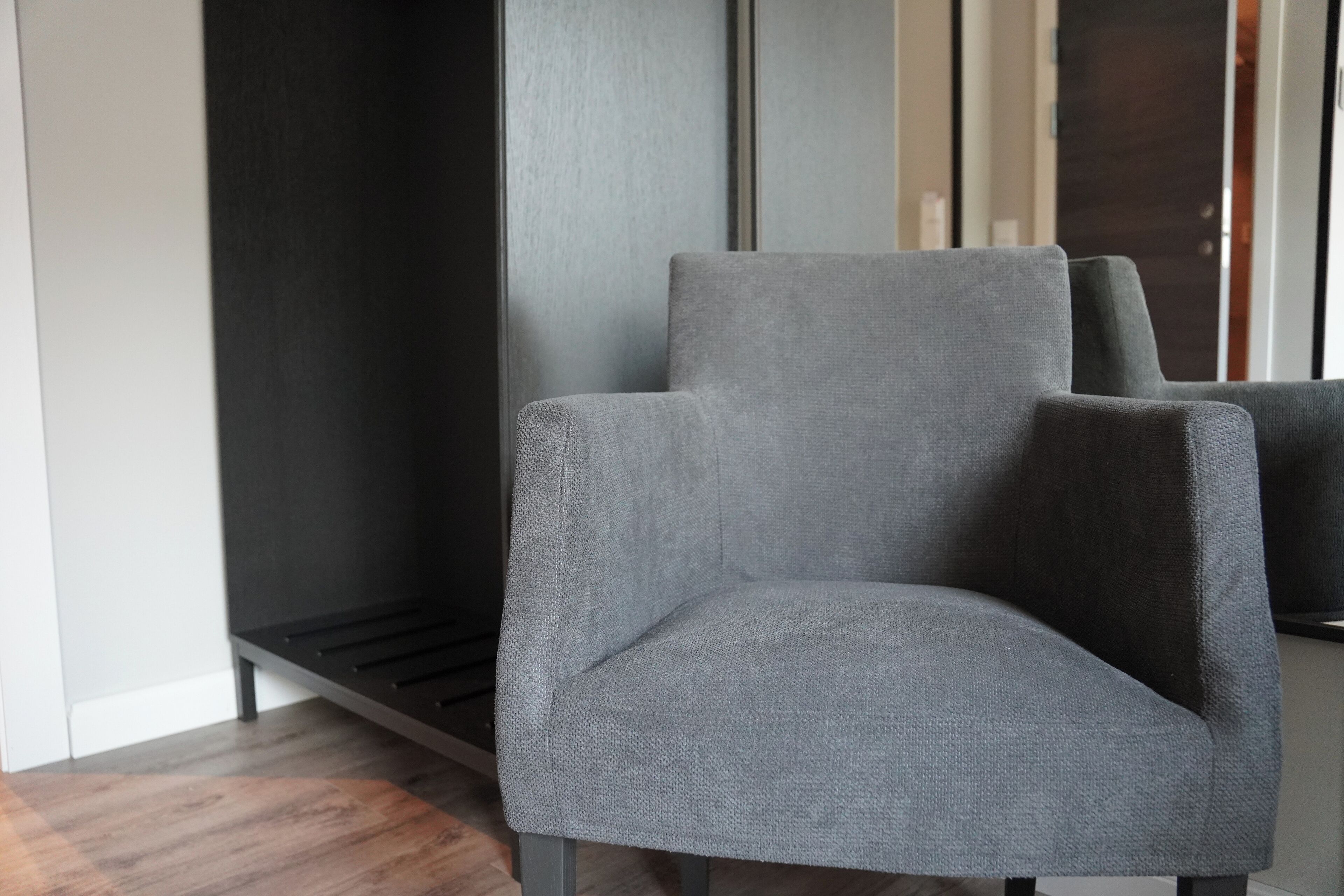 Un sillón tapizado gris con respaldo alto, ubicado en un rincón de una habitación con suelo oscuro y un amplio ventanal.