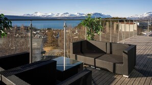Una terraza con mobiliario de mimbre, barandilla de vidrio y vistas a un lago y montañas.