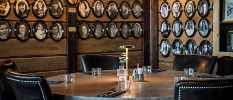 Un restaurant avec un mur recouvert de photos encadrées, une table ronde entourée de chaises et une petite table sur laquelle se trouvent une lampe et des verres.