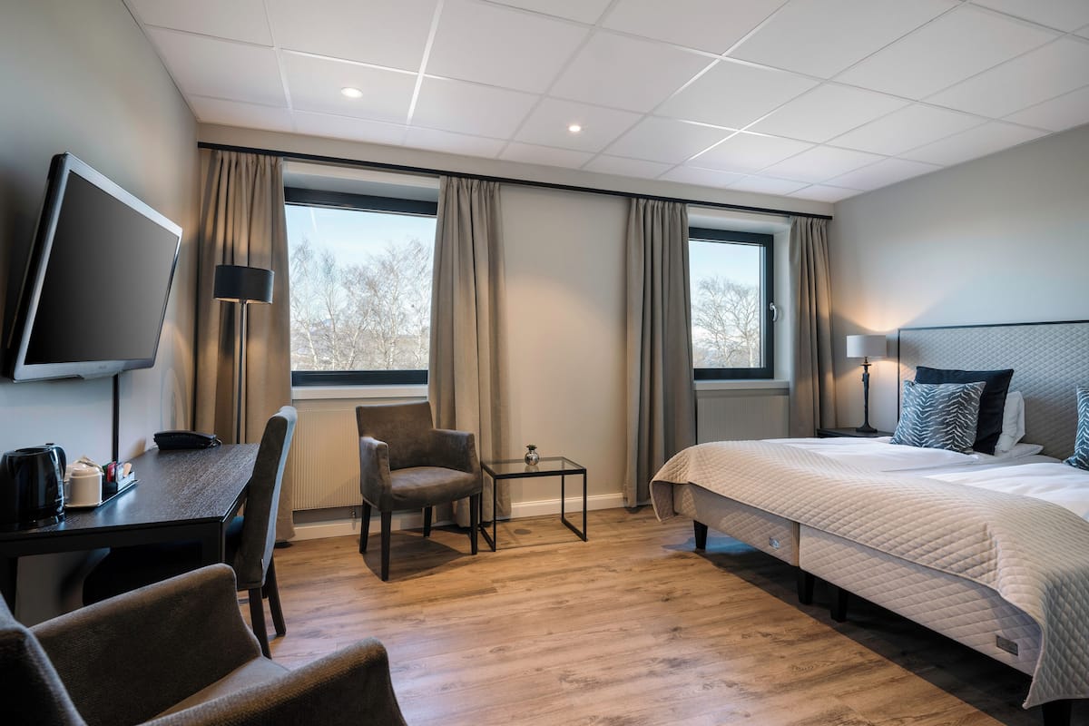 A hotel room with a bed, a desk, a chair, a TV, and two windows with curtains.