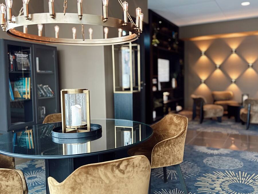 En modern hotellobby med ett matbord med glasskiva, bekväma sittplatser, en dekorativ vägg och en kristallkrona.