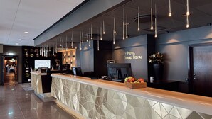 Zona de recepción de hotel con diseño moderno, que incluye un mostrador de recepción, un televisor montado en la pared y un cartel que dice 'Quality Hotel Grand Royal'.