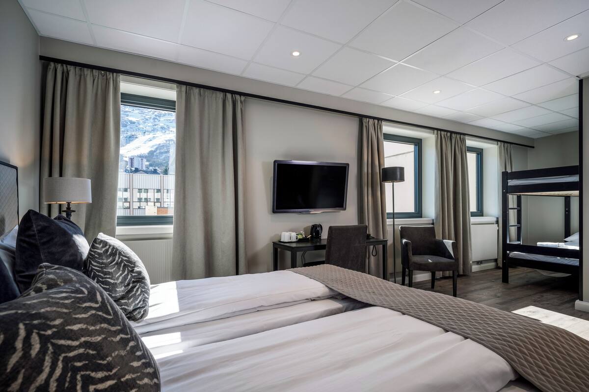 A hotel room with a large bed, a desk with a chair, a TV, and a bunk bed.
