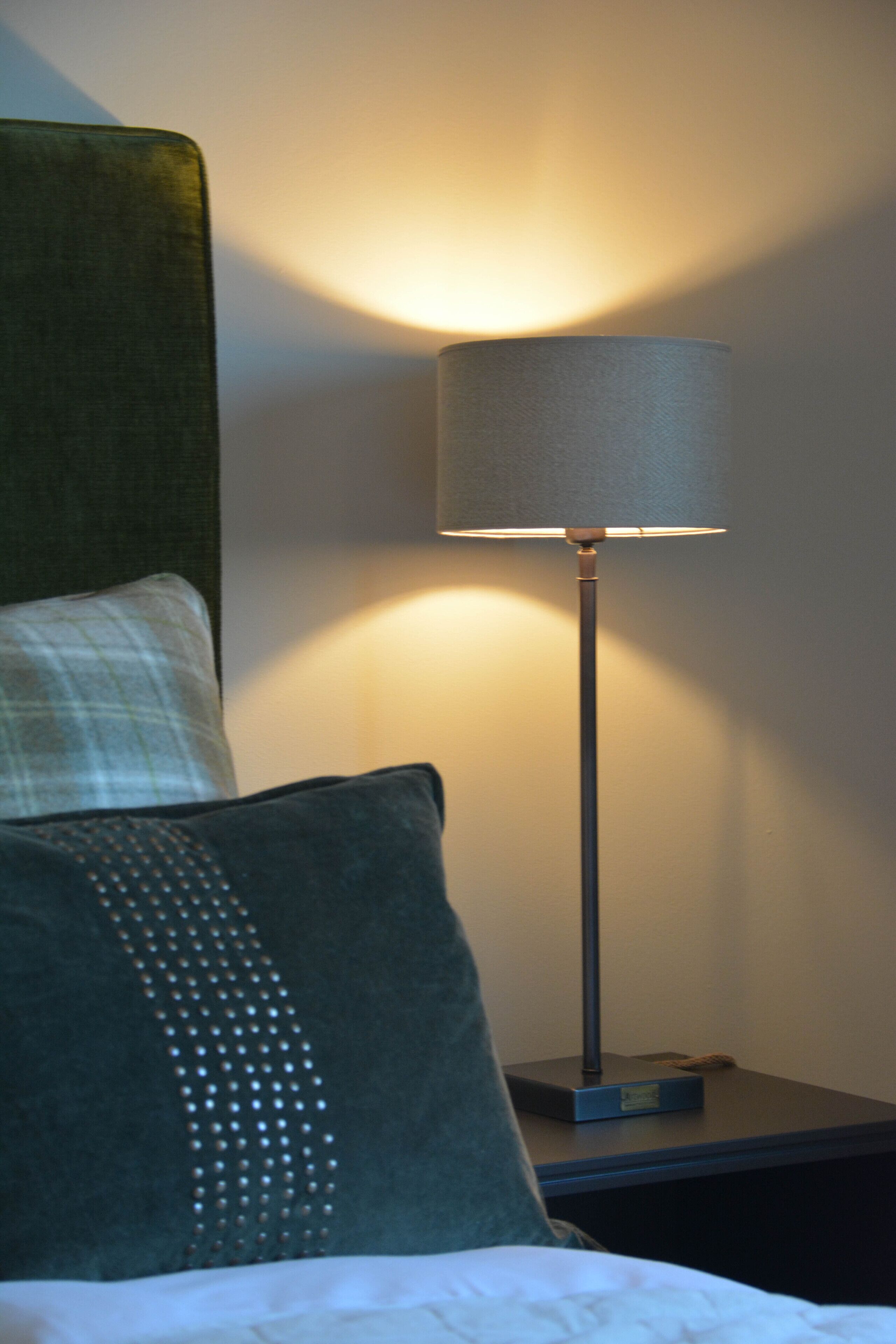 A bed with a green headboard and a pillow with a dotted pattern, a bedside table with a lamp, and a wall with a shadow cast by the lamp.
