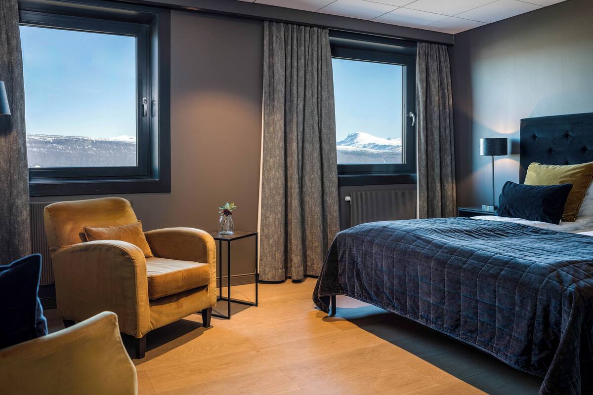 A hotel room with a bed, a chair, a small table with a vase, and two windows with curtains.