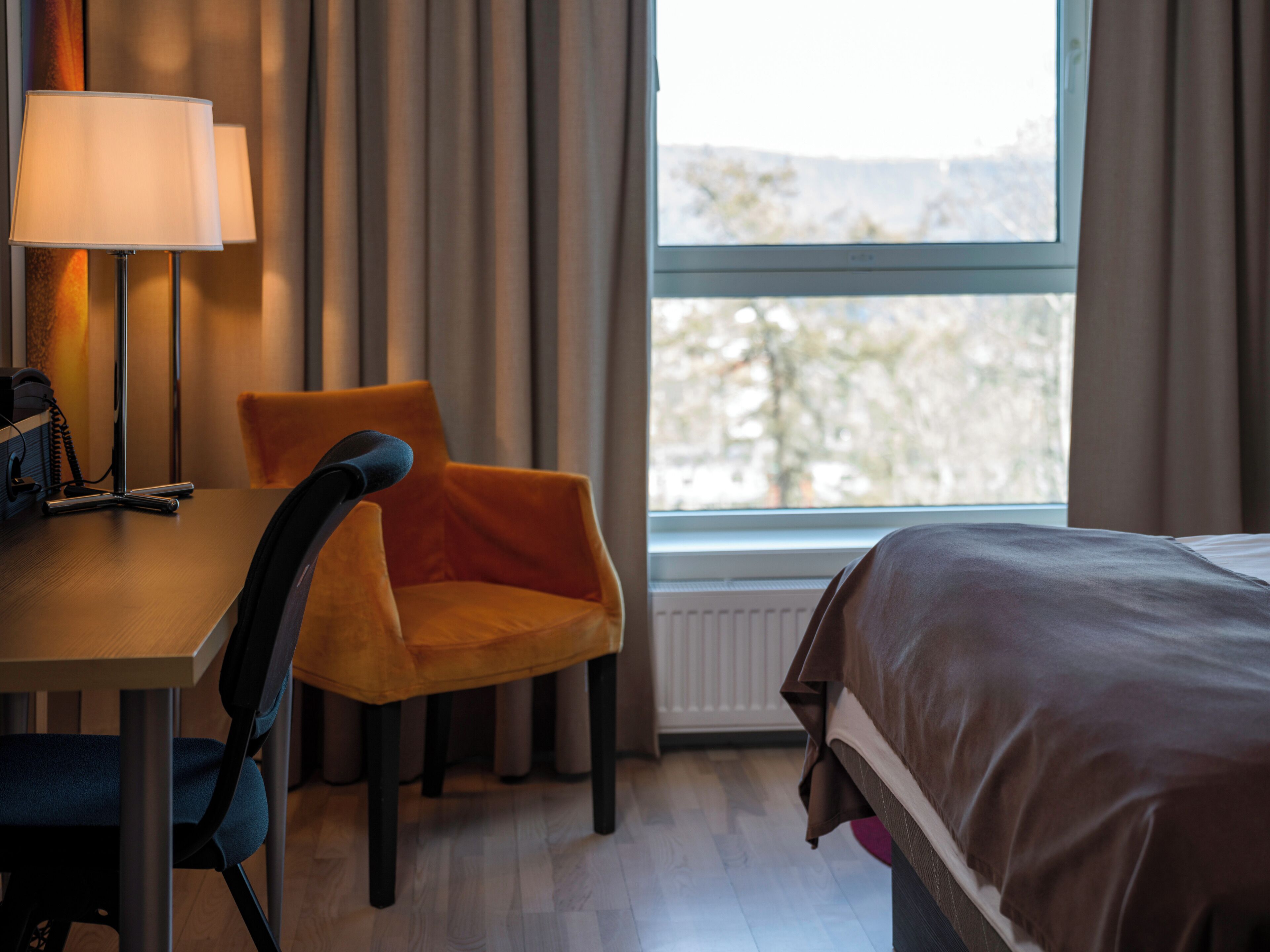 A hotel room with a bed, a desk with a lamp, a chair, and a view of the outdoors through a window.