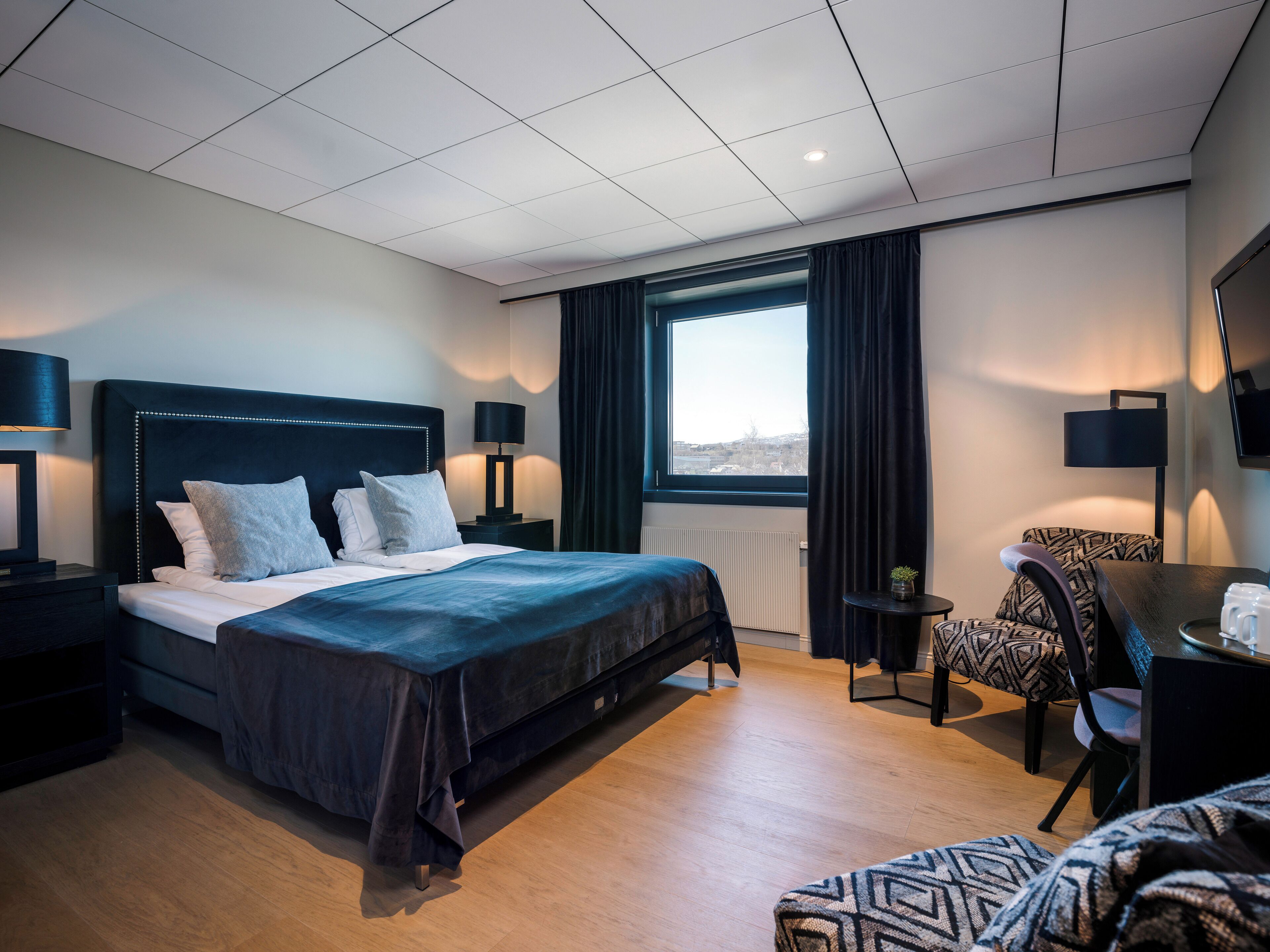 A modern hotel room with a large bed, a desk, a chair, and a view of the city through the window.