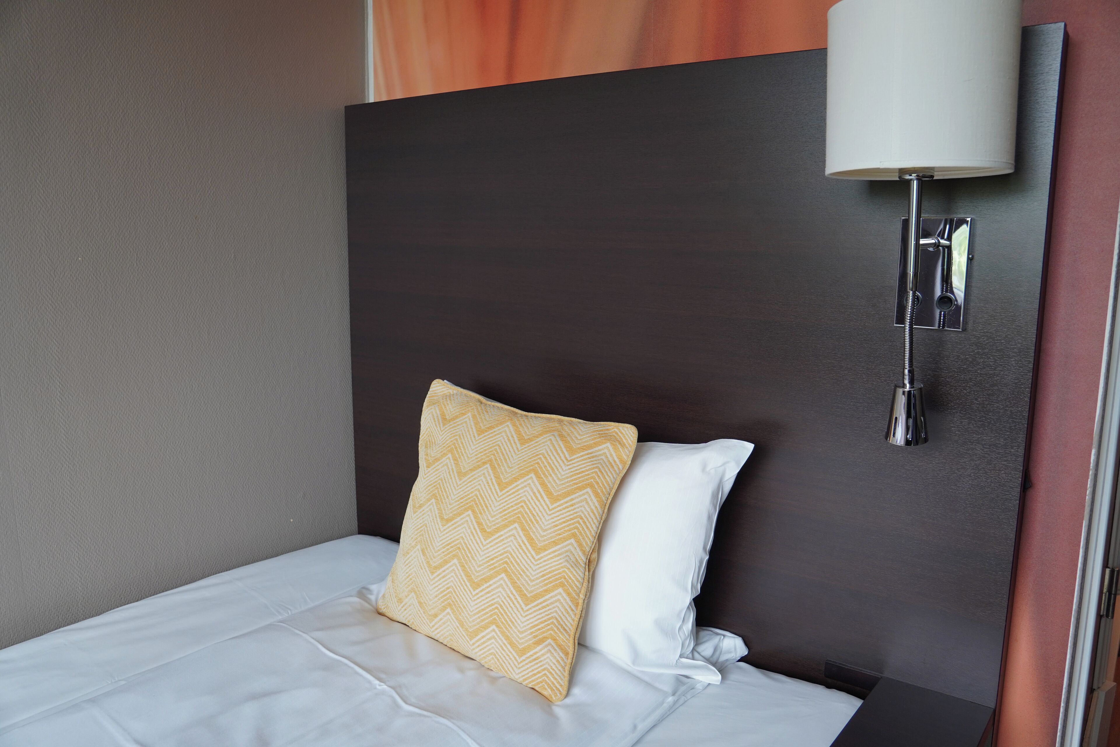 Habitación de hotel con una cama, un cojín estampado y otro liso, además de una lámpara fijada a la pared.