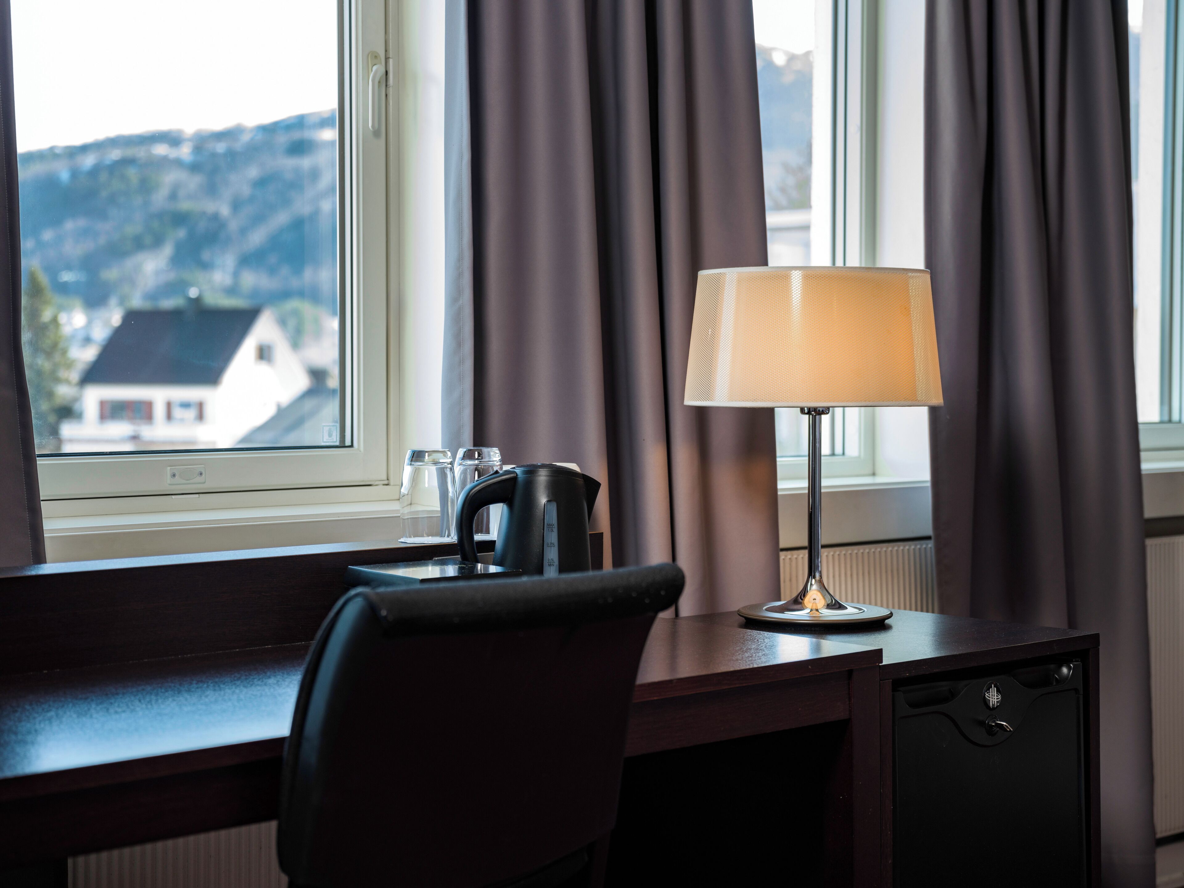 A hotel room with a desk, a lamp, a kettle, and a view of a house through a window.