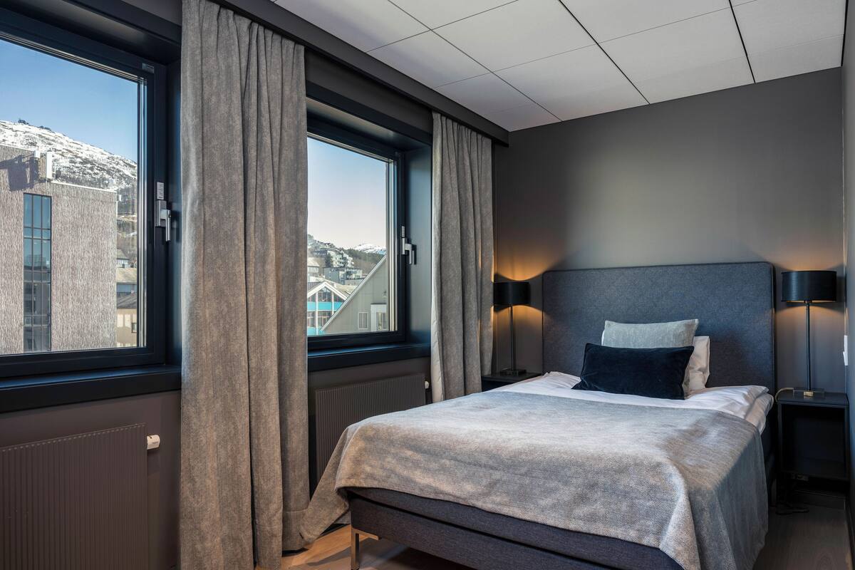 A modern hotel room with a large window, a bed with a grey headboard, and two bedside tables with lamps.