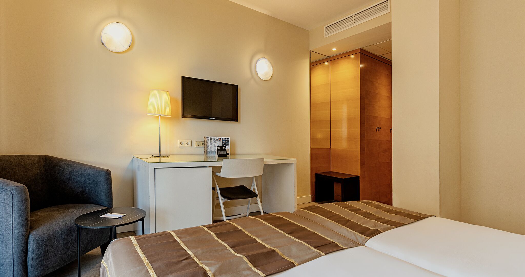 A hotel room with a bed, a desk, a chair, a lamp, and a television.