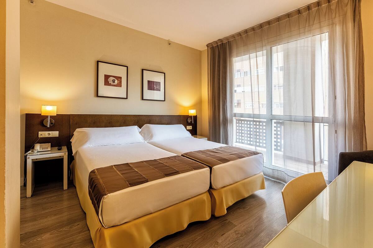 Habitación de hotel con cama doble, mesitas de noche con lámparas, una pequeña mesa y vistas al exterior a través de amplias ventanas.