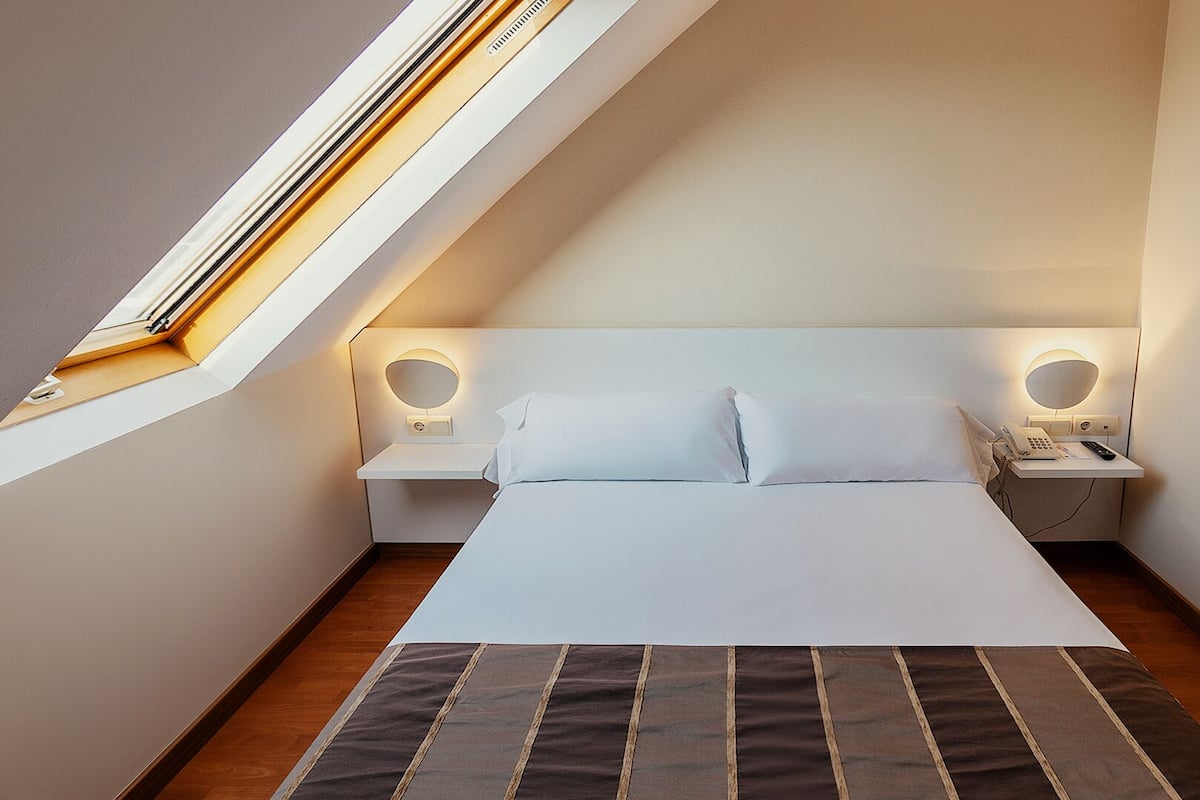 Un dormitorio con tragaluz, cama con ropa de cama blanca, dos mesitas de noche con lámparas y un teléfono pequeño en la mesita de noche.