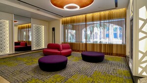 Une chambre d’hôtel moderne avec un tapis à motifs, des fauteuils rouges et violets, de grandes fenêtres avec des rideaux et un panneau mural décoratif.
