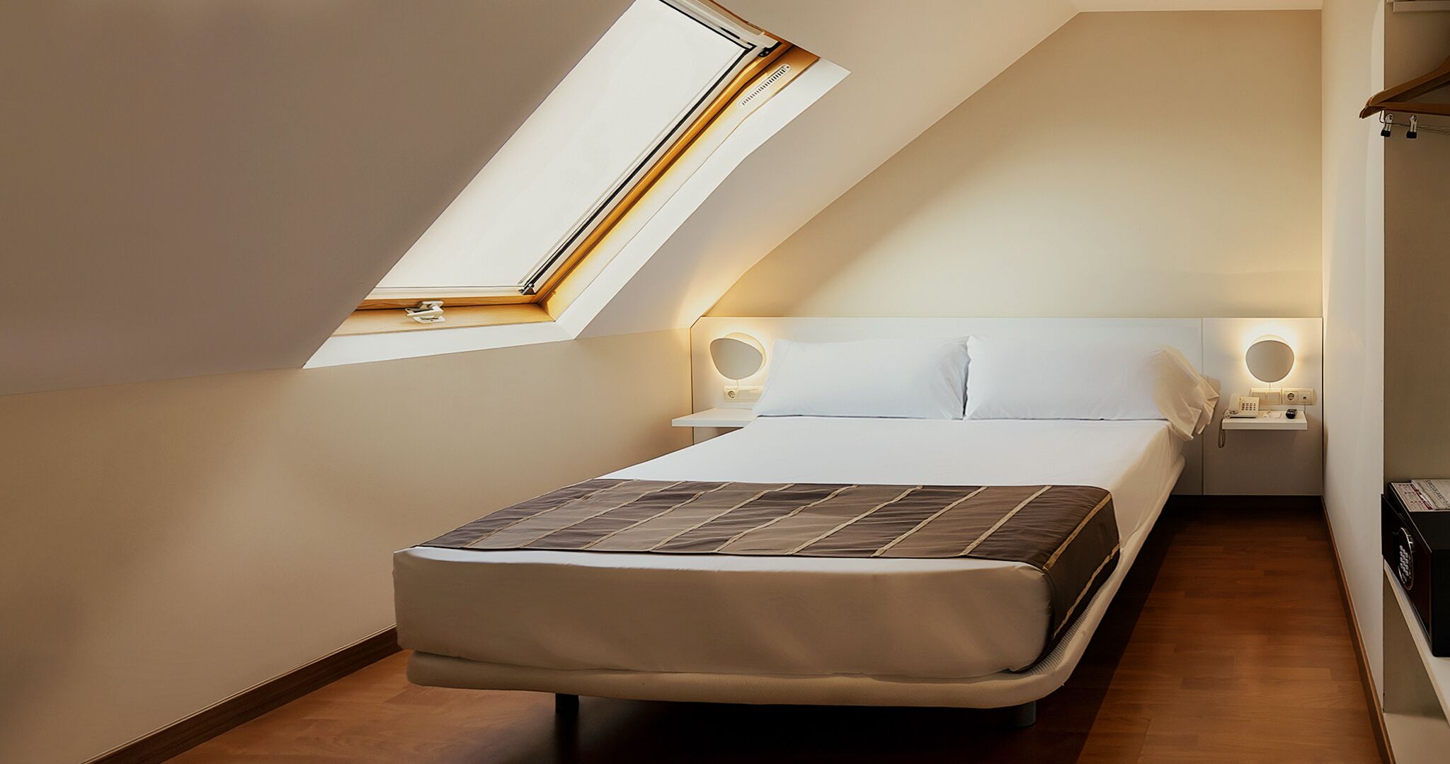 A small, modern bedroom with a sloped ceiling and a skylight, featuring a bed, bedside tables, and a small bedside lamp.