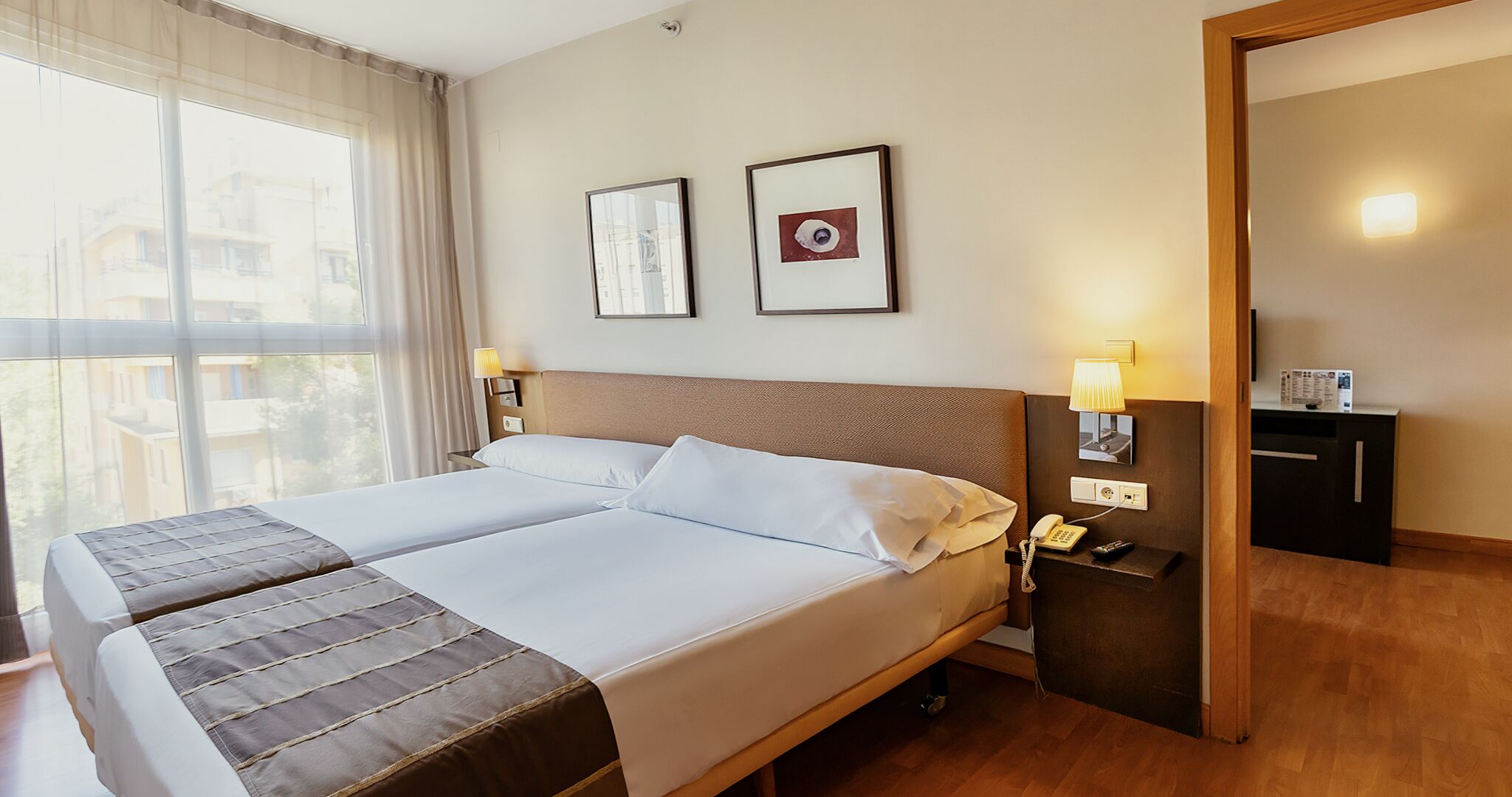 Cama doble con cabecera, lámparas de noche, mesita de noche con teléfono y control remoto, y vista al exterior a través de amplias ventanas.