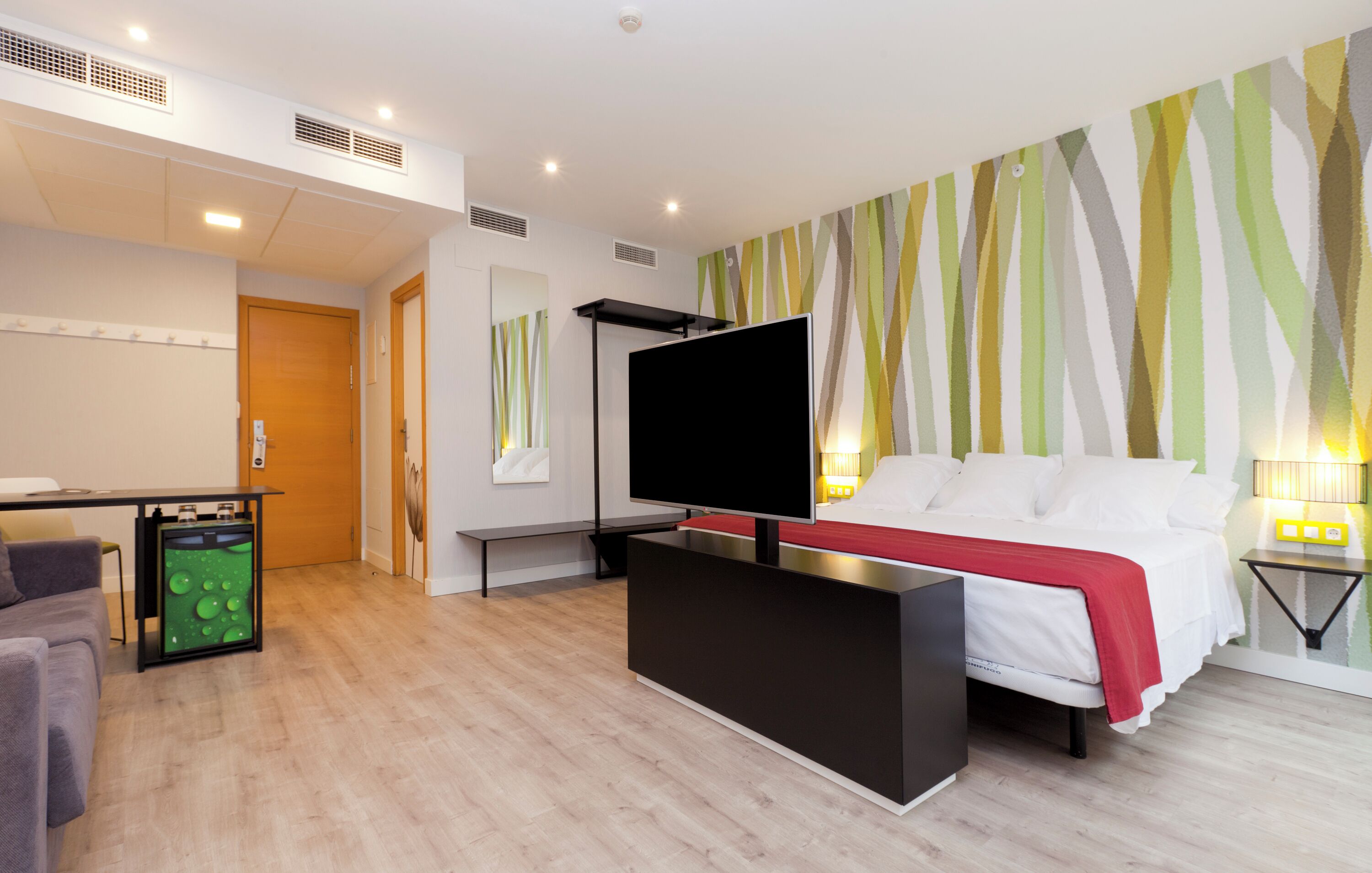 A modern hotel room with a bed, TV, desk, and a wall with a striped pattern.