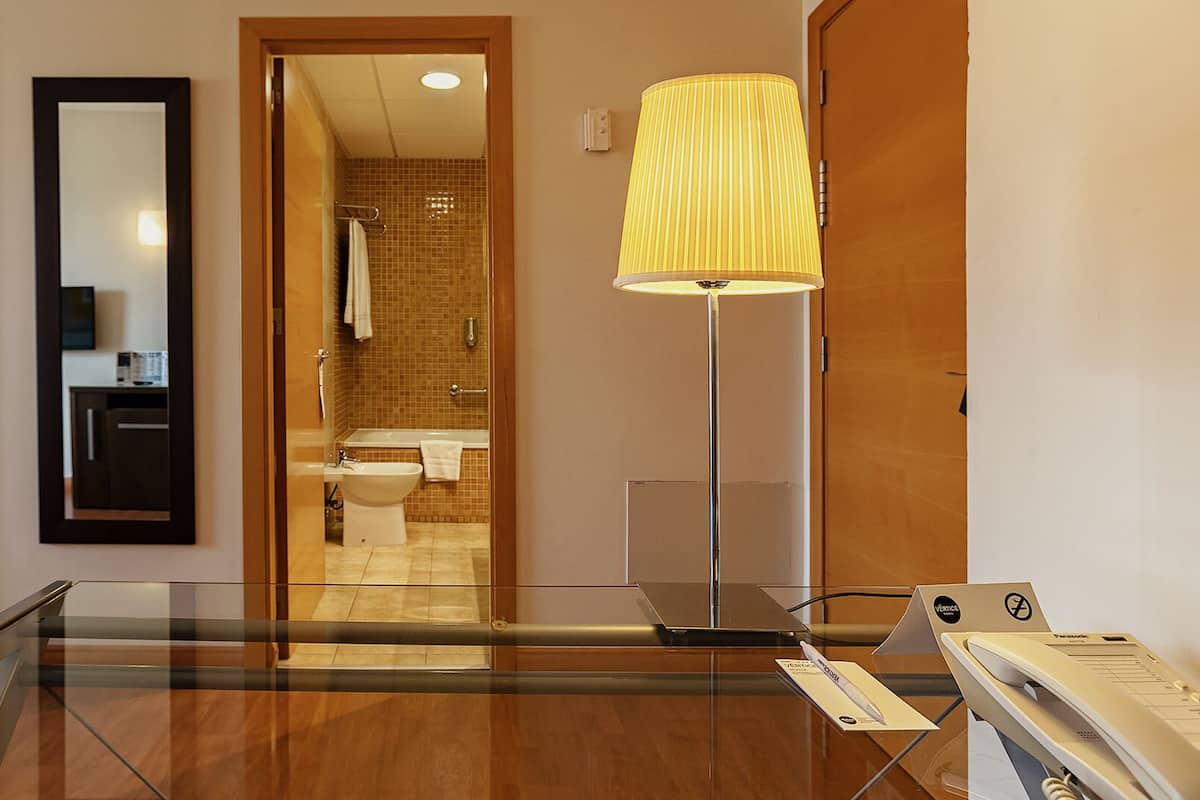 Habitación de hotel con cama, espejo, baño con lavamanos, inodoro y ducha. Hay una mesa de vidrio con una lámpara y un teléfono.