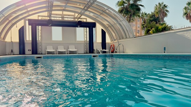 Une piscine intérieure à l’eau claire, entourée de chaises longues blanches et dotée d’une structure de plafond en verre.