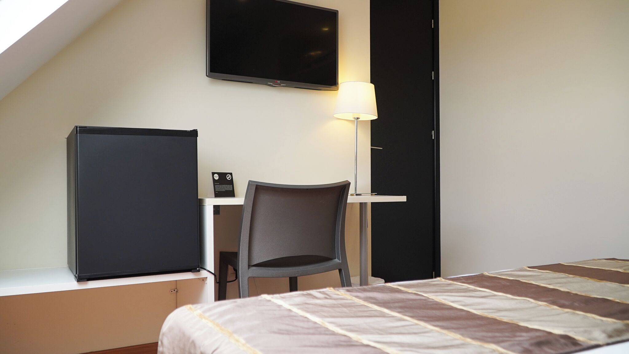 A small, modern hotel room with a bed, a desk with a chair, a flat-screen TV, and a lamp.