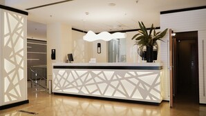 Un comptoir d’accueil moderne, orné de motifs géométriques, agrémenté d’une plante en pot et offrant une vue sur l’extérieur à travers les stores.