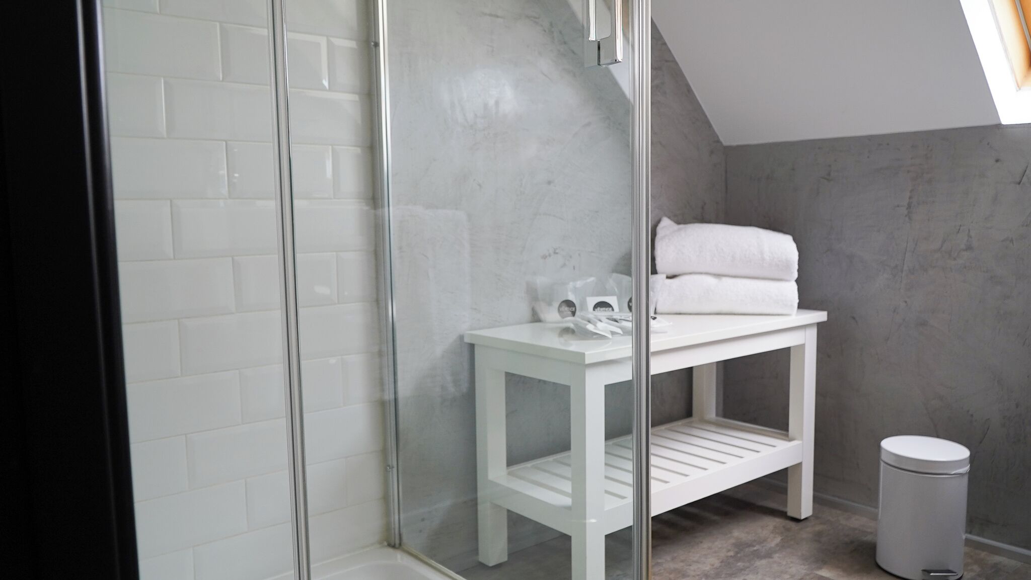 A modern bathroom with a glass shower enclosure, a white vanity with a towel rack, and a trash can.