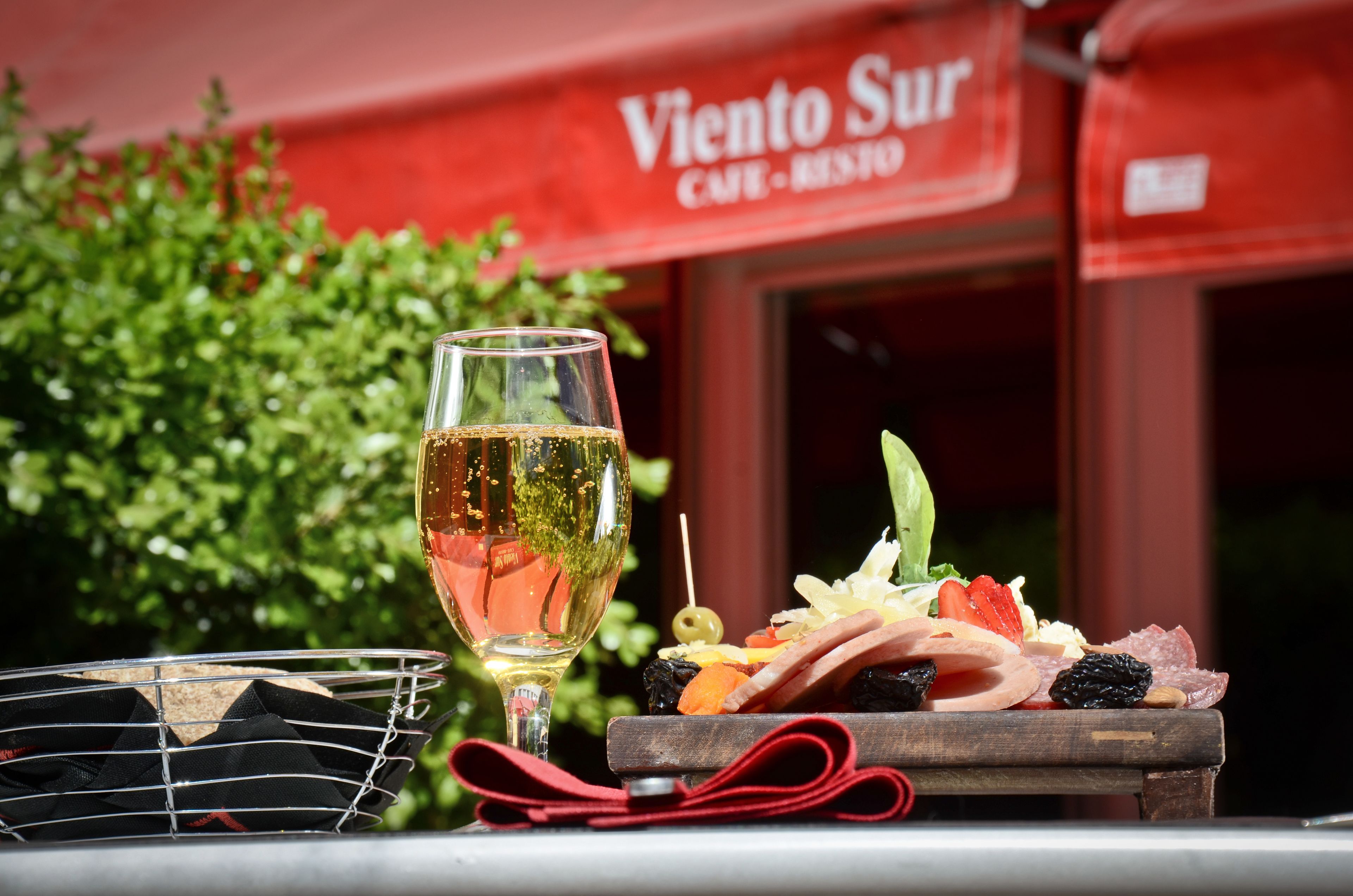 A glass of wine and a charcuterie board on a table outside a cafe named "Viento Sur".