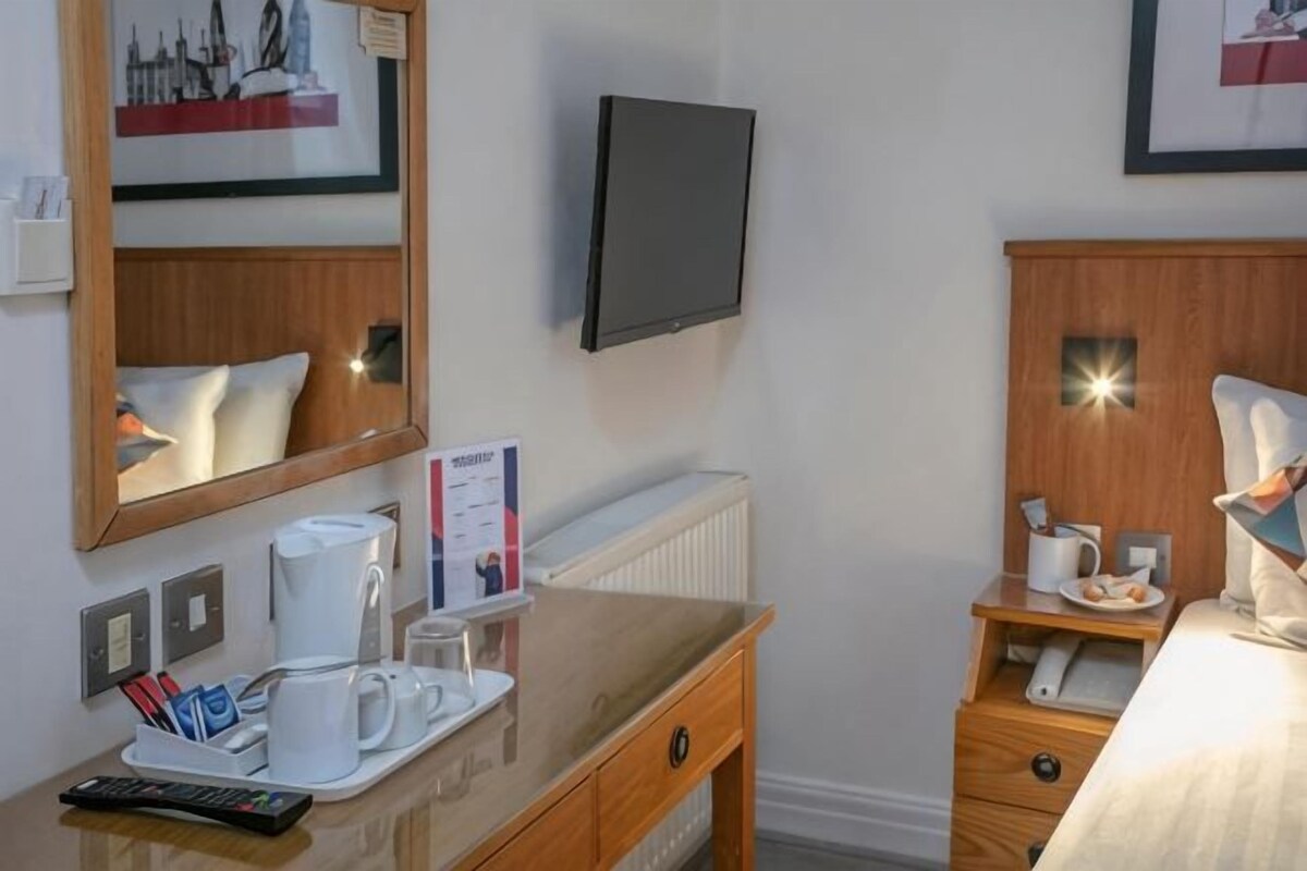 A hotel room with a wooden dresser, a bedside table with a lamp, a television mounted on the wall, and a framed picture above the dresser.