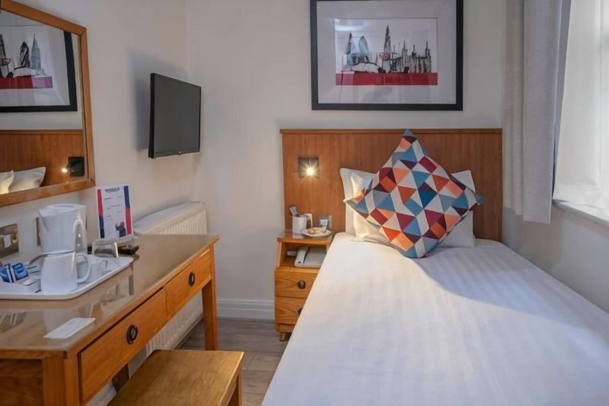 A hotel room with a bed, a bedside table with a lamp, a desk with a chair, a mirror, and a framed picture on the wall.