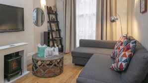 A living room with a flat-screen TV, a fireplace, a grey sofa, a patterned pillow, and a small round table with shopping bags. - Queensway Hotel, Sure Hotel Collection by Best Western (London)