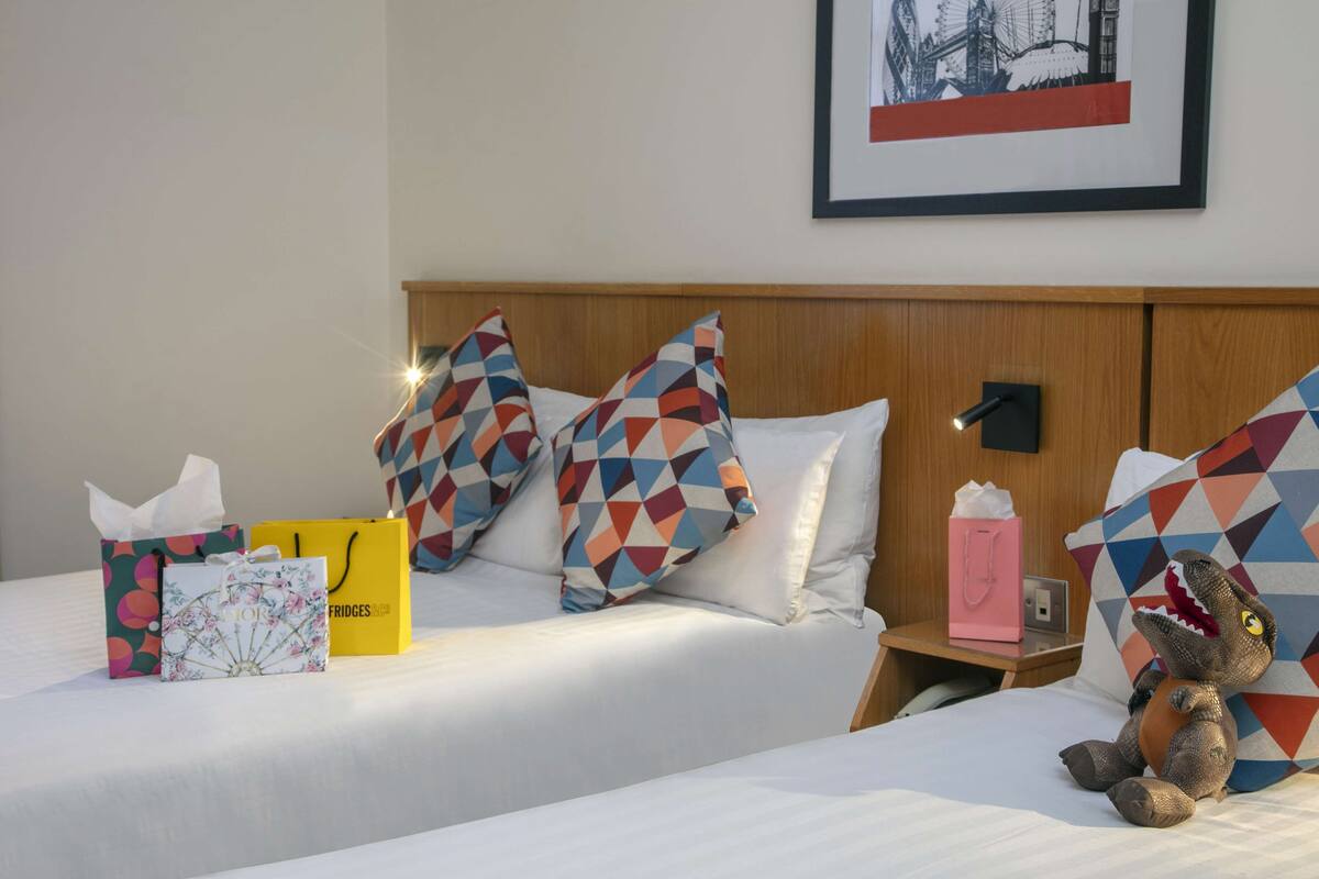 A hotel room with two beds, colorful geometric pillows, a dinosaur plush toy, and a framed picture on the wall.