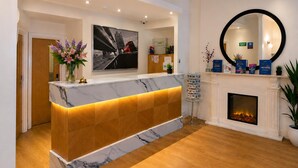 A reception desk with a marble top, a wooden panel, and a lit fireplace. - Queensway Hotel, Sure Hotel Collection by Best Western (London)